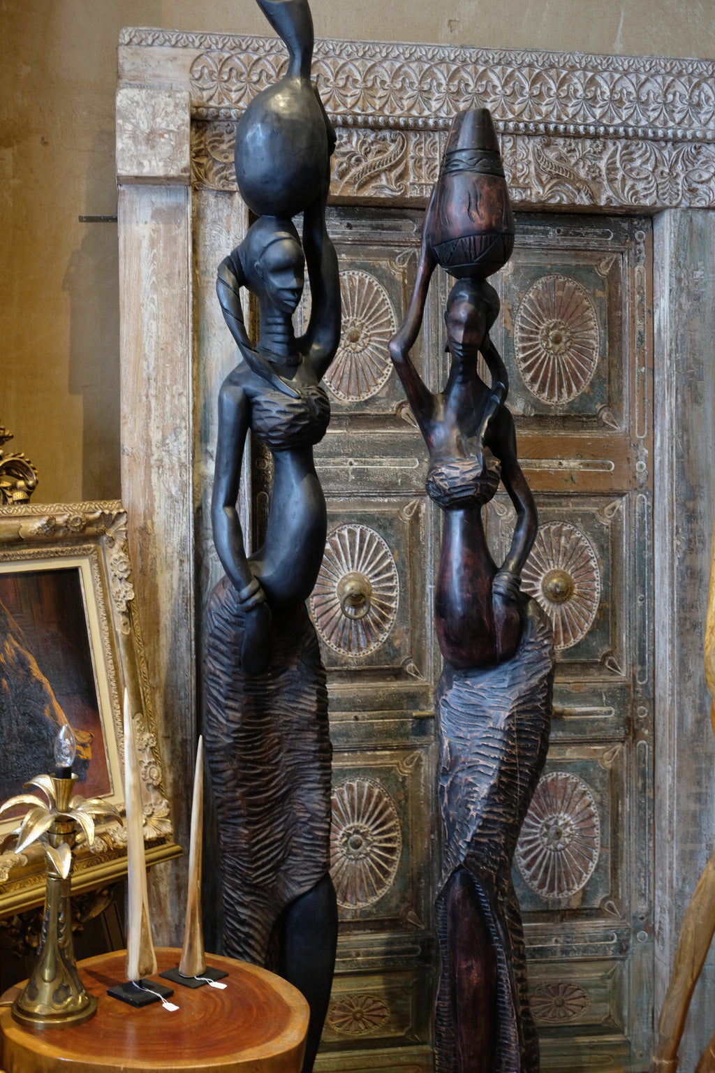 Two dark carved African-style female statues standing in front of an ornate carved wooden door, with one statue holding a tall black vessel on its head.