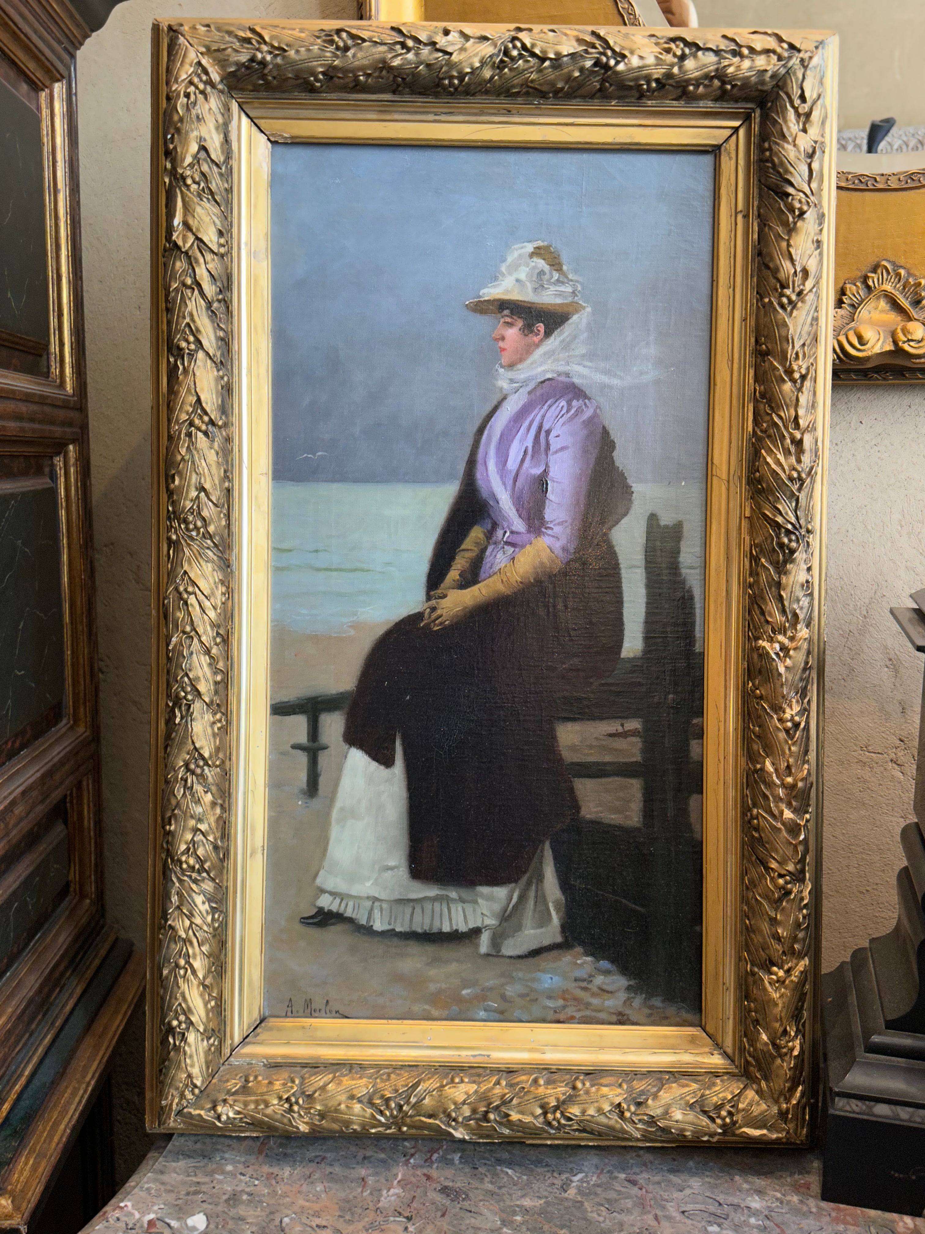 A framed oil painting of a woman seated by the sea, shown in an ornate gold frame. The painting depicts the woman in a lavender and dark dress with a white hat, set against a muted blue-gray sky and sea background.