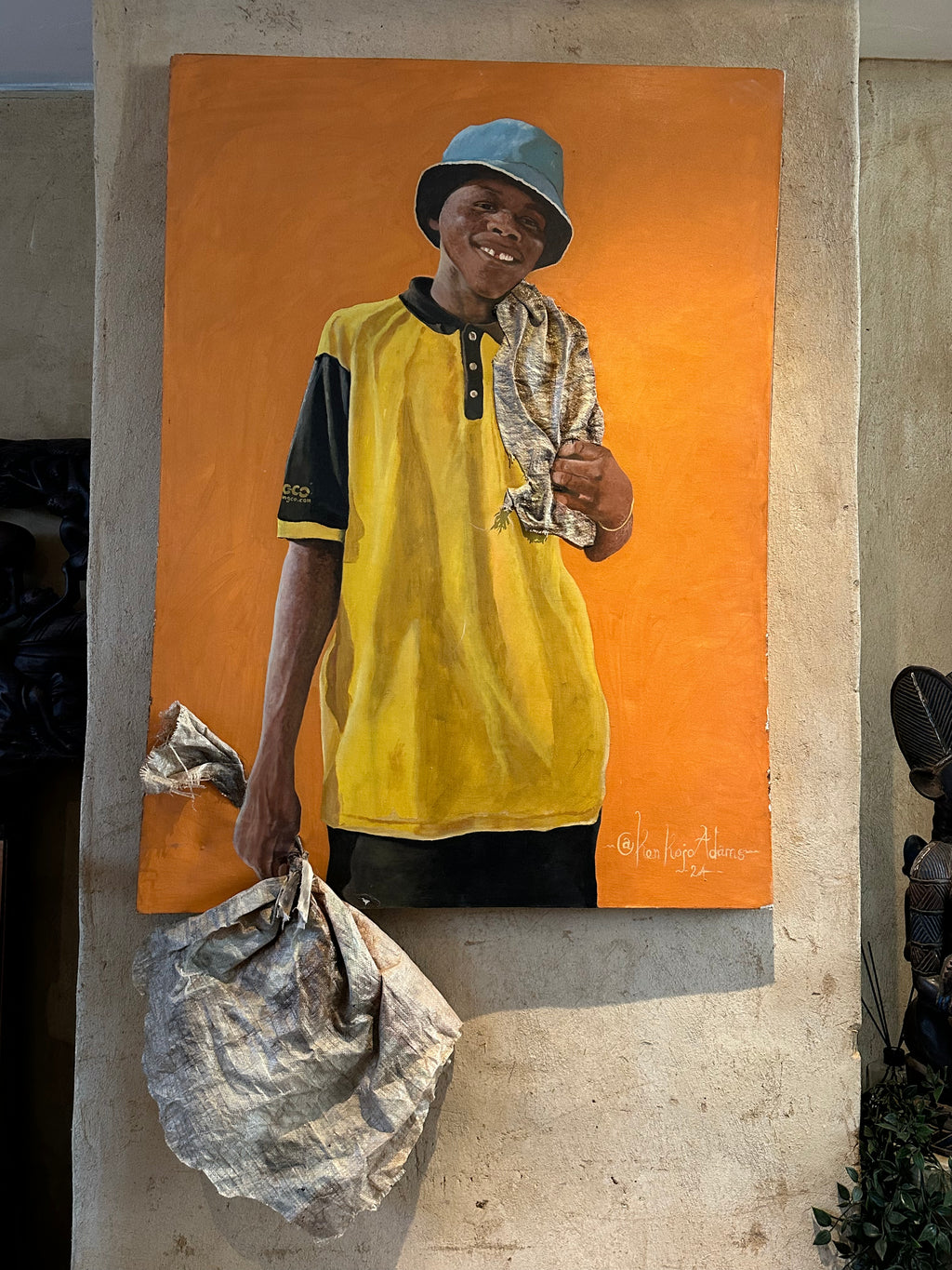 A large vertical canvas painting, depicting a smiling man in a yellow and black shirt with a blue hat holding a crumpled bag against a bright orange background.