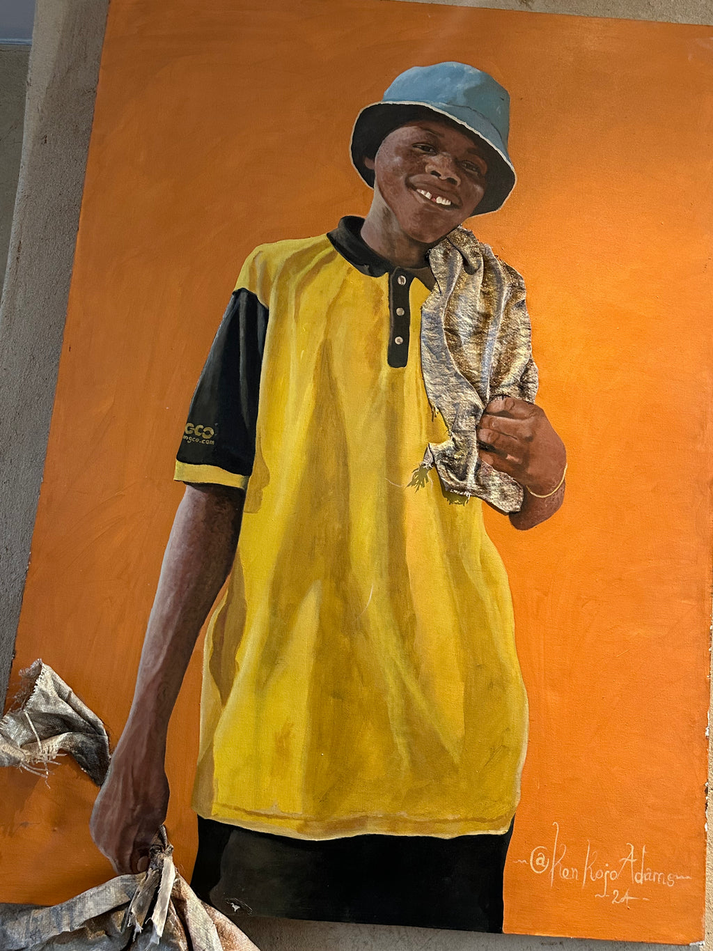 A painted portrait of a smiling man wearing a blue bucket hat, a yellow polo shirt with black sleeves, and holding a patterned cloth, set against a large orange background. The artwork is signed in the lower right corner.