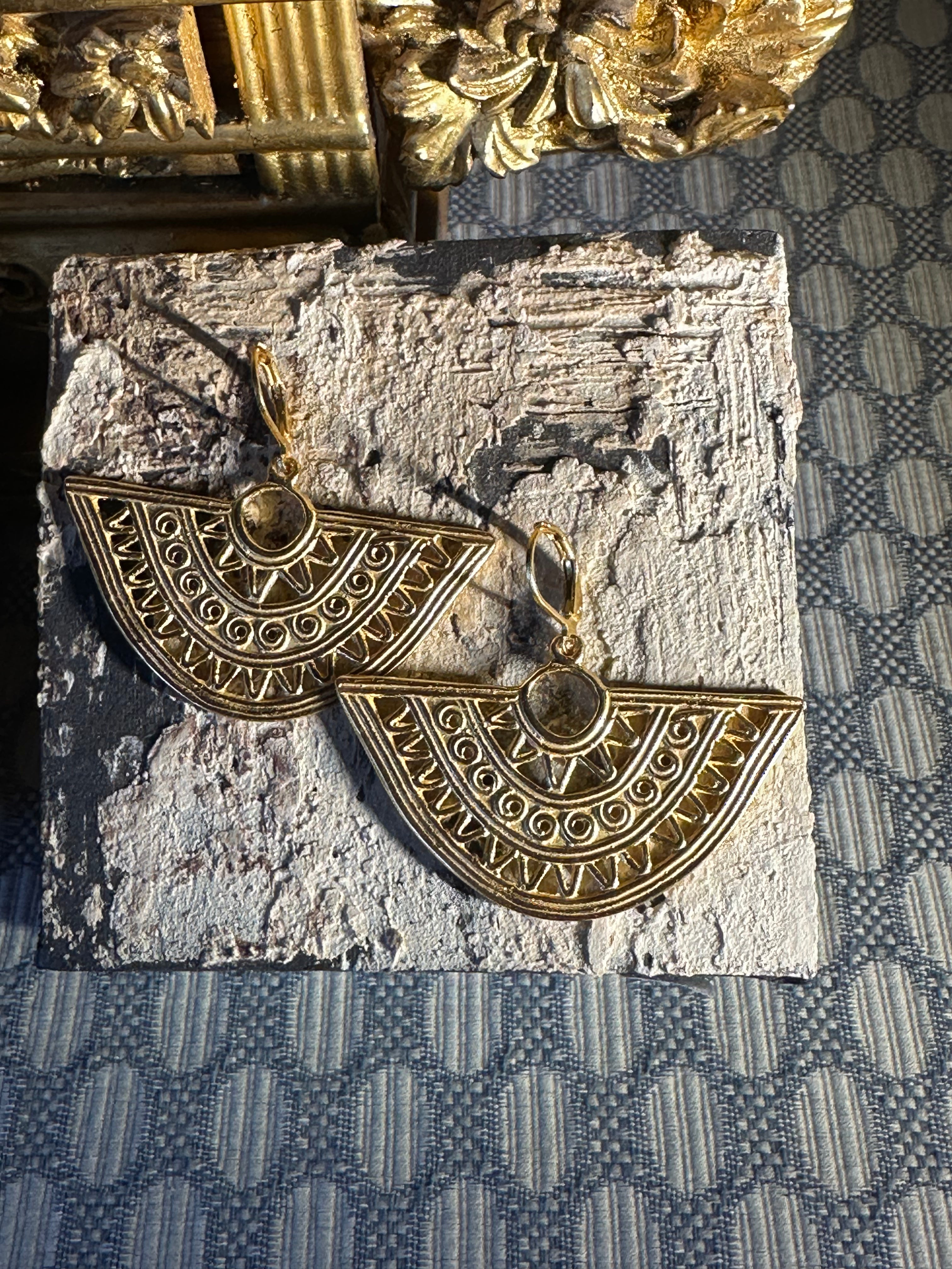 A pair of gold-toned geometric dangle earrings with intricate filigree detailing, displayed on a textured stone slab over a blue patterned fabric background.