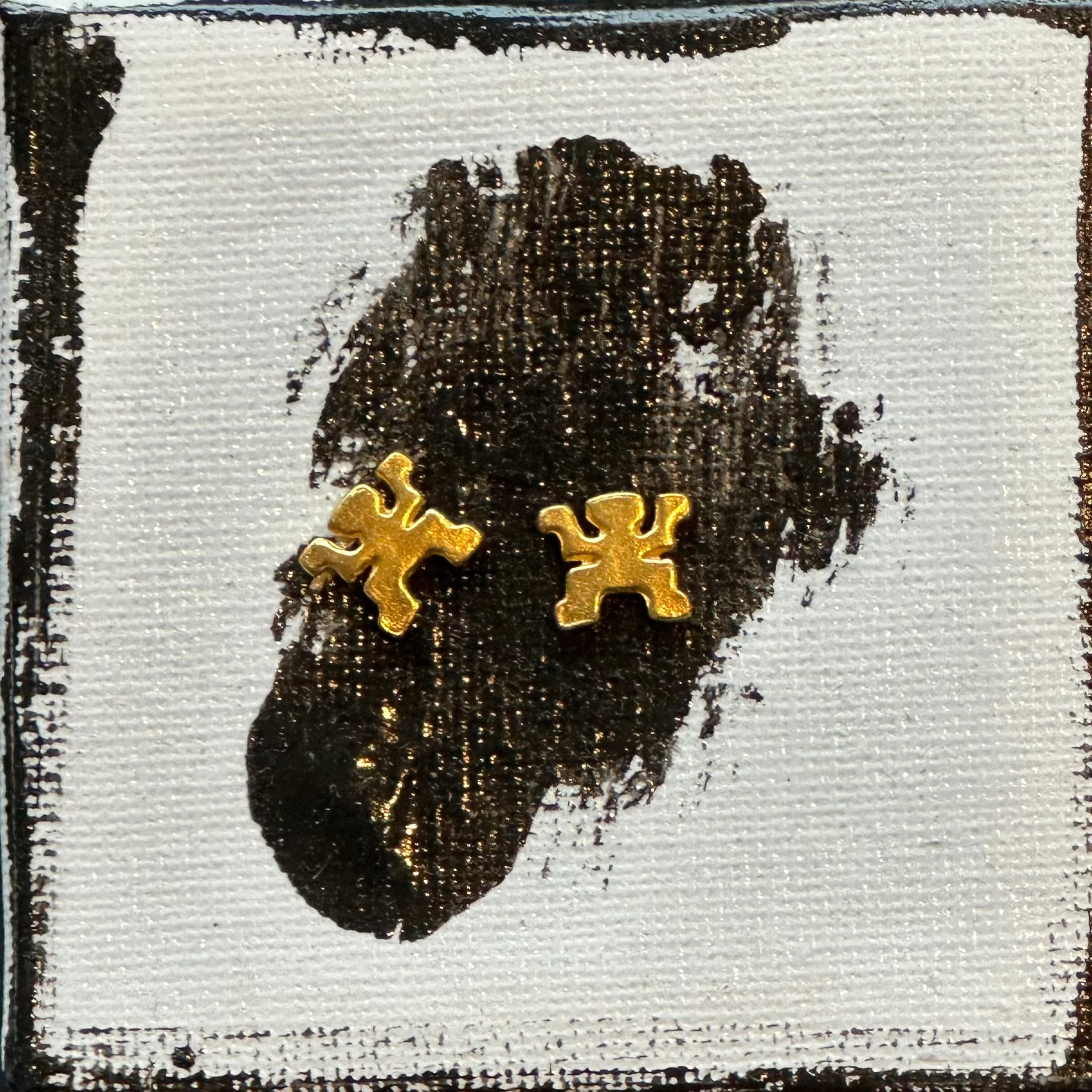 A pair of gold-colored puzzle-piece stud earrings displayed on a square canvas print, which is set against a dark gray marble surface.