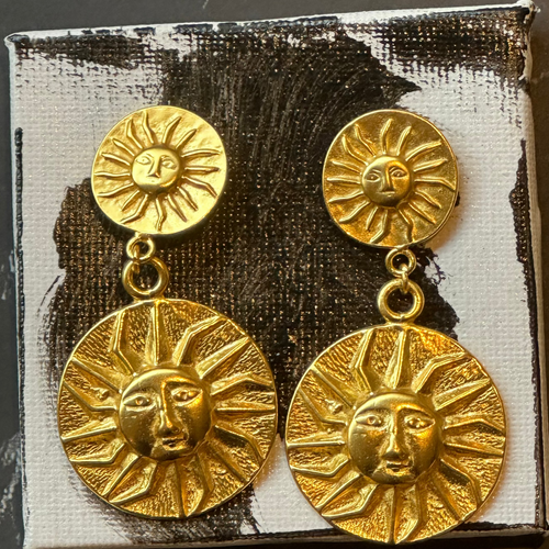 A pair of gold-toned dangle earrings with sun-face disc charms, displayed on a small white and black textured box against a dark gray surface.
