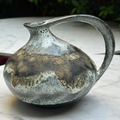 A handmade ceramic pitcher with a rounded body, a curved handle, and a small spout, featuring a textured glaze with brown, gray, and white mottled patterns, resting on a white marble surface.