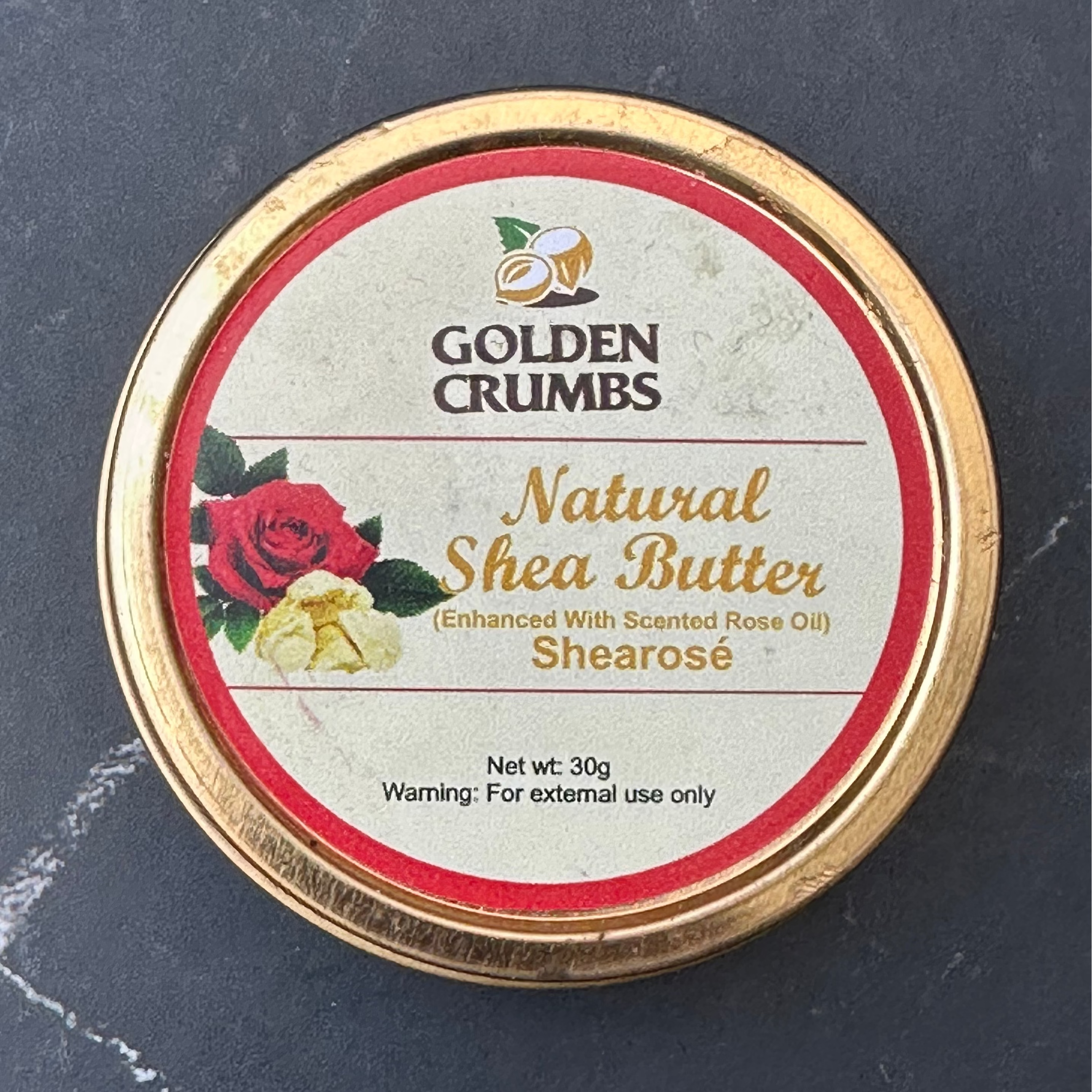 A round tin of Golden Crumbs Natural Shea Butter, featuring a gold-colored metal rim and a cream-colored label with red and gold text, set against a dark slate background.