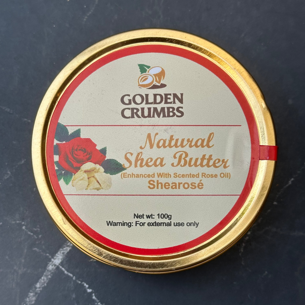 A round tin of Golden Crumbs Natural Shea Butter (Shearosé) with a gold-colored metal rim and a cream-colored label featuring a red rose and golden coconut logo, set against a dark gray marble background.
