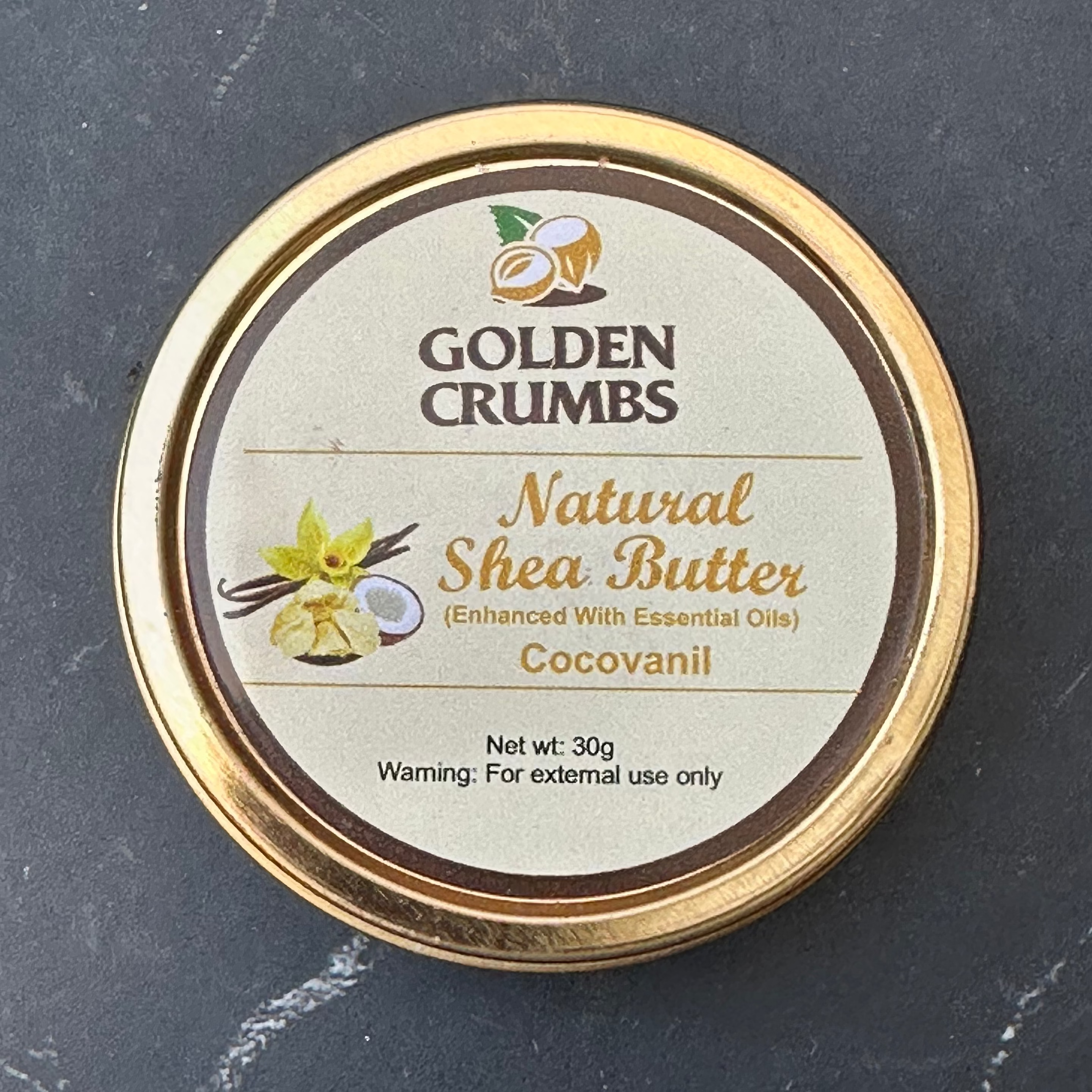 A round tin of Golden Crumbs Natural Shea Butter (Cocovanil) with a gold-colored rim and a beige label, placed on a dark gray stone surface.