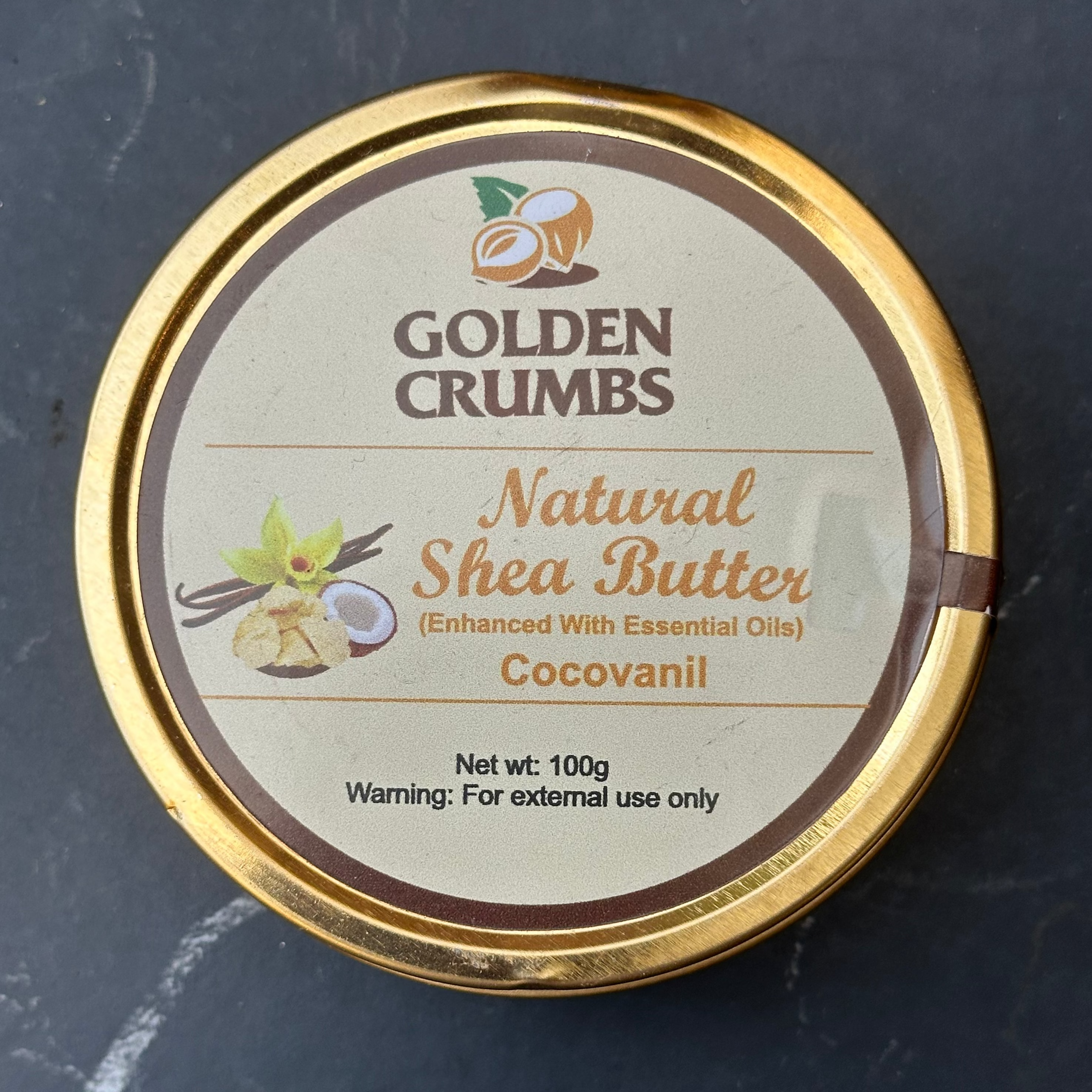 A round gold metal tin of Golden Crumbs Natural Shea Butter (Cocovanil) on a dark gray marble surface. The label features a cream background with brown and gold text and illustrations of shea nuts and a coconut.
