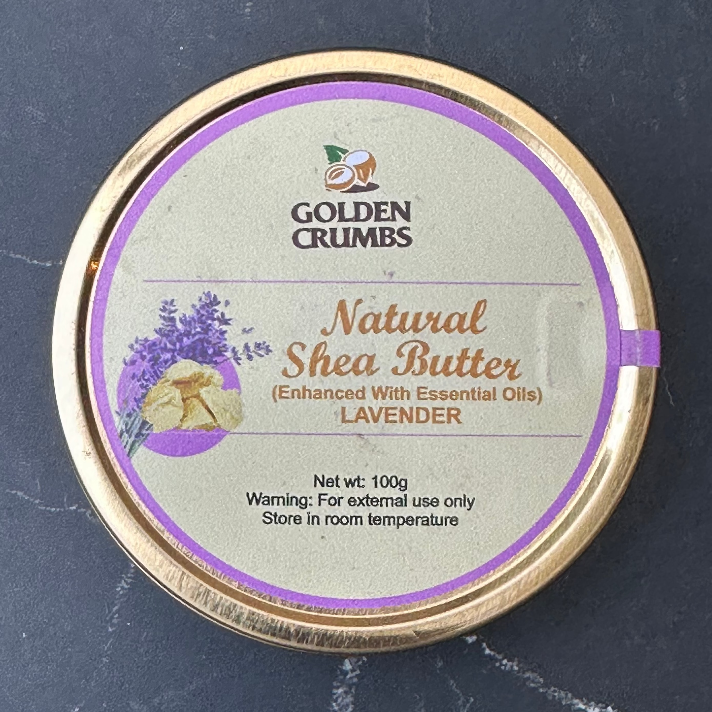 A round tin of Golden Crumbs Natural Shea Butter in Lavender scent, featuring a light beige label with purple accents and a gold-colored rim, set against a dark slate background.