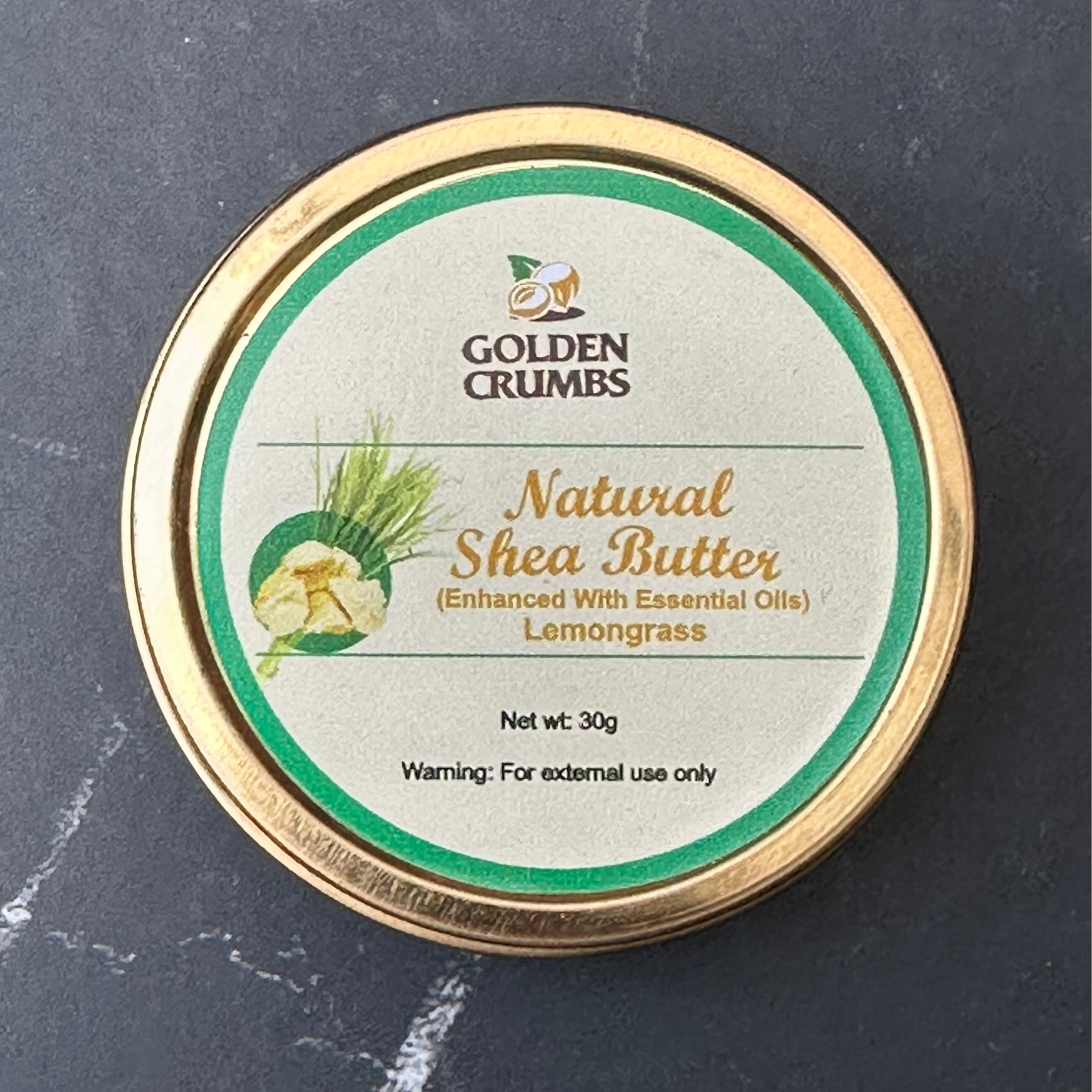 A round tin of Golden Crumbs Natural Shea Butter (Enhanced with Essential Oils) Lemongrass, set on a dark gray marble surface.