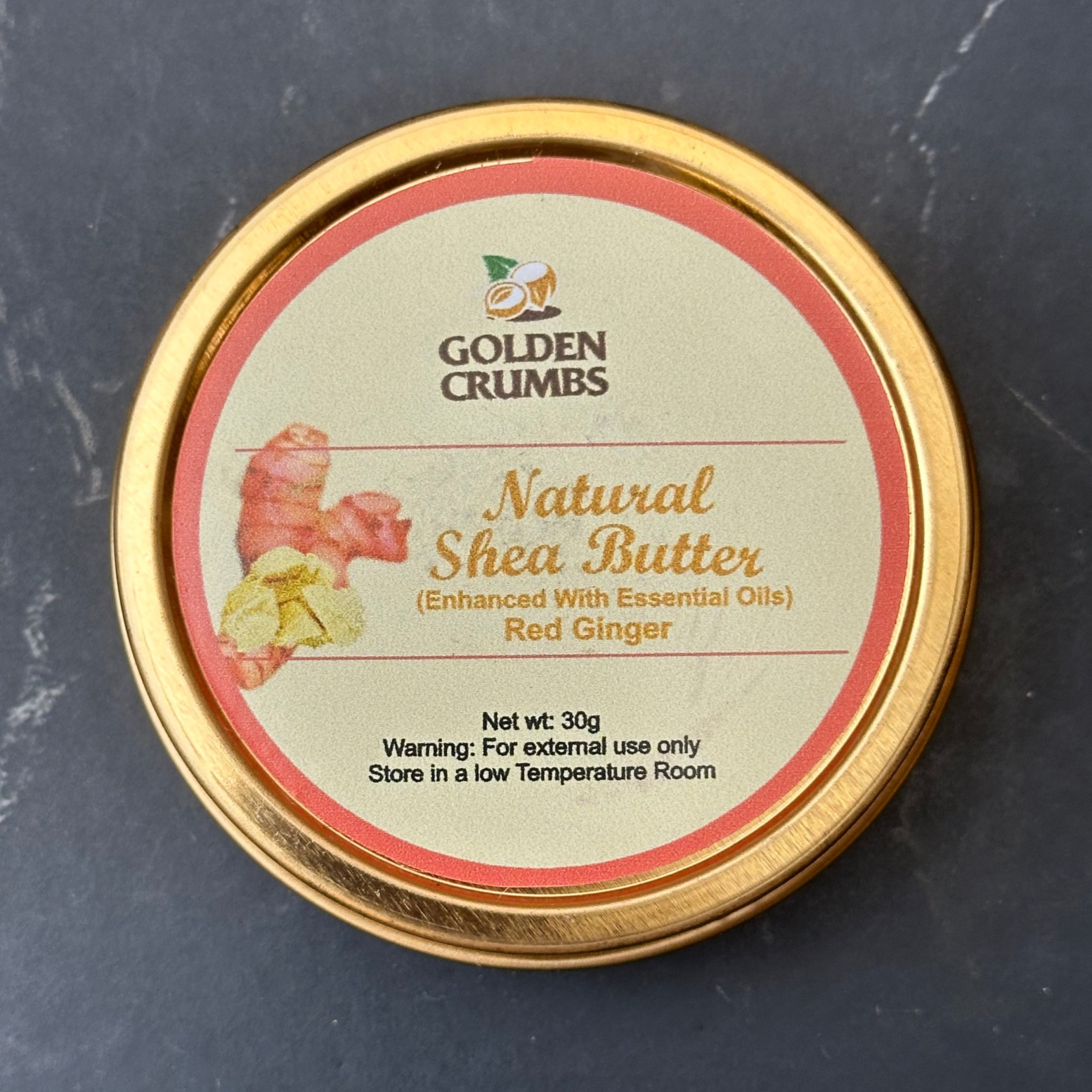 A round gold metal tin of Golden Crumbs Natural Shea Butter (Enhanced with Essential Oils) Red Ginger, shown on a dark gray marble surface.