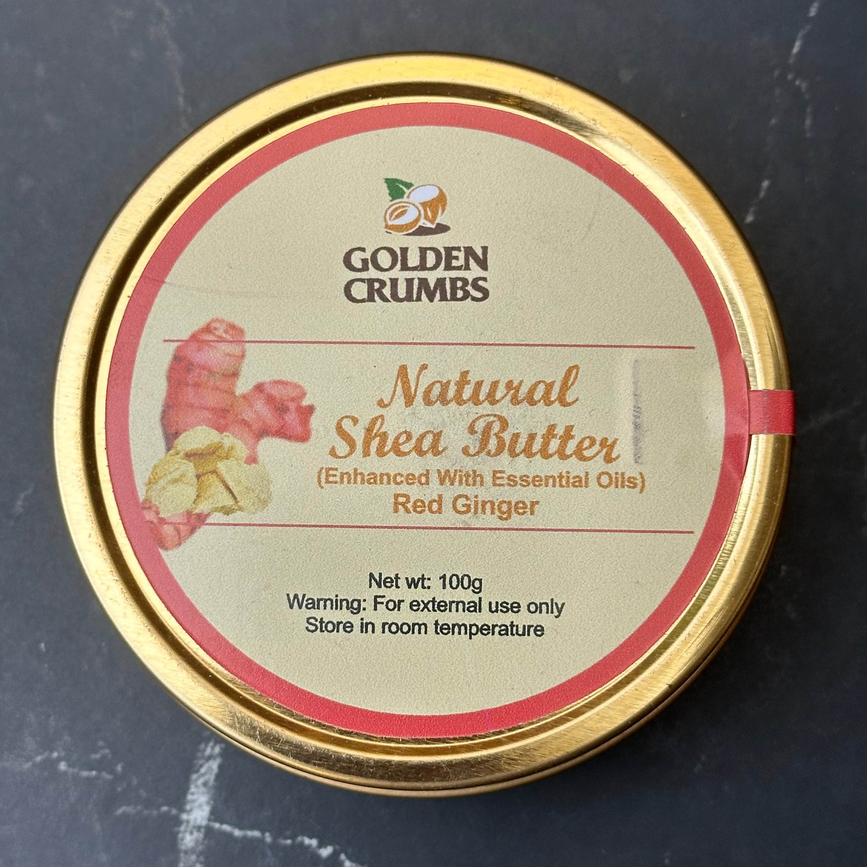 A round gold metal tin of Golden Crumbs Natural Shea Butter (Red Ginger variant) on a dark gray marble surface.