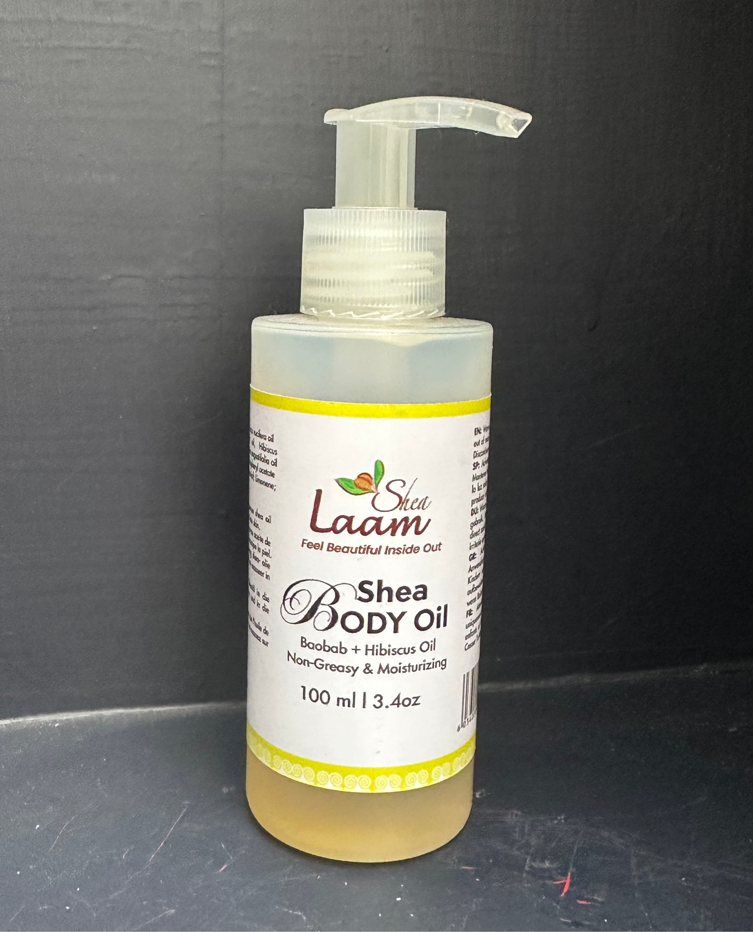 A bottle of Shea Body Oil by Shea Laam, featuring a clear plastic pump dispenser and a white label with yellow accents, containing a golden-yellow oil, set against a dark gray background.